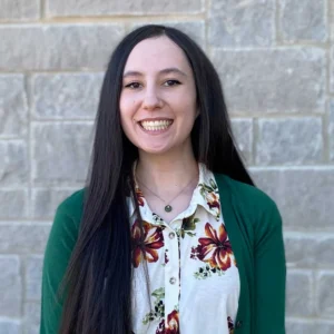 Young woman with long dark hair wearing a green cardigan over a floral patterned white top, smiling at camera against brick background