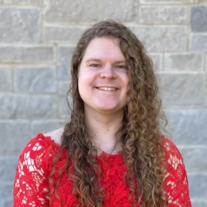 Woman with long curly brown hair smiling at camera, wearing red patterned sweater against brick wall background