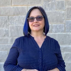 Woman with dark blue hair and glasses smiling at camera wearing dark blue polka dot top against brick wall