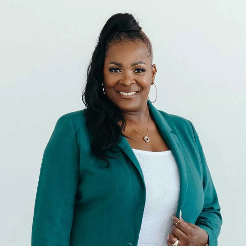 Woman with long black hair in a teal green blazer over white top, wearing a necklace and smiling at camera against white background