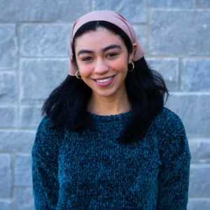 Young woman wearing pink headband and teal sweater with black fur collar smiling at camera