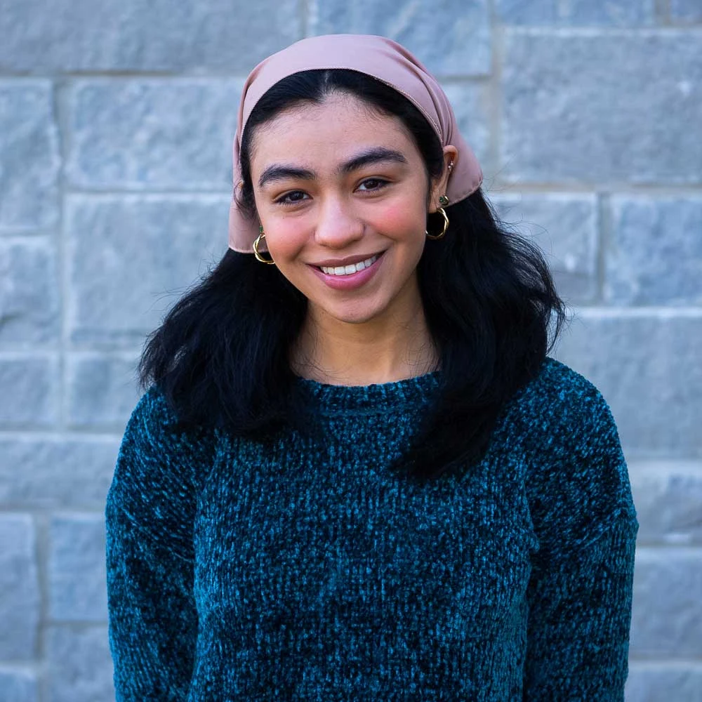 Young woman wearing pink headband and teal sweater with black fur collar smiling at camera