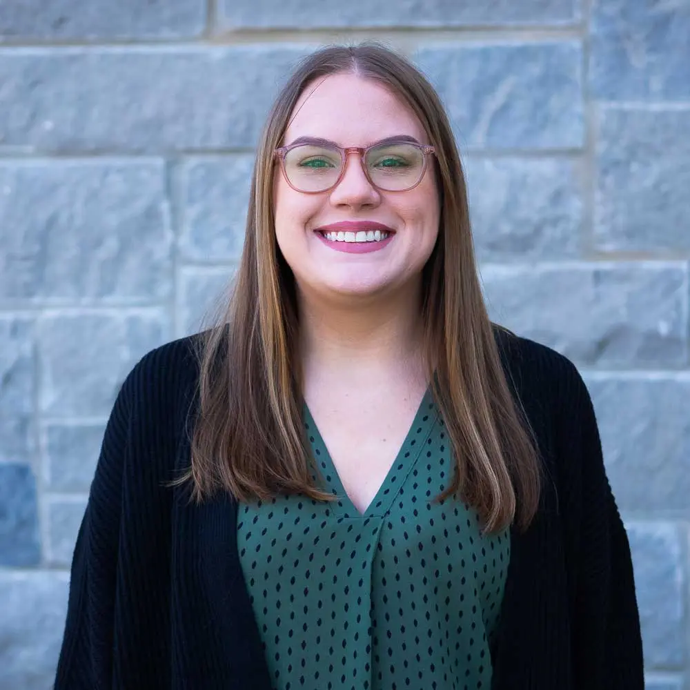 Woman with long brown hair and green glasses smiling at camera wearing dark top against brick wall background