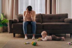 young mother sitting on couch with her head in her hands and in need of postpartum depression treatment while baby plays on floor