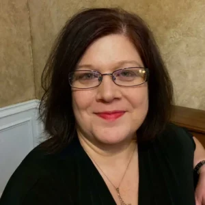 A woman with shoulder-length dark hair and glasses smiling at the camera, wearing black clothing, photographed indoors against a neutral background