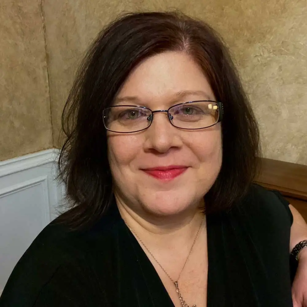 A woman with shoulder-length dark hair and glasses smiling at the camera, wearing black clothing, photographed indoors against a neutral background
