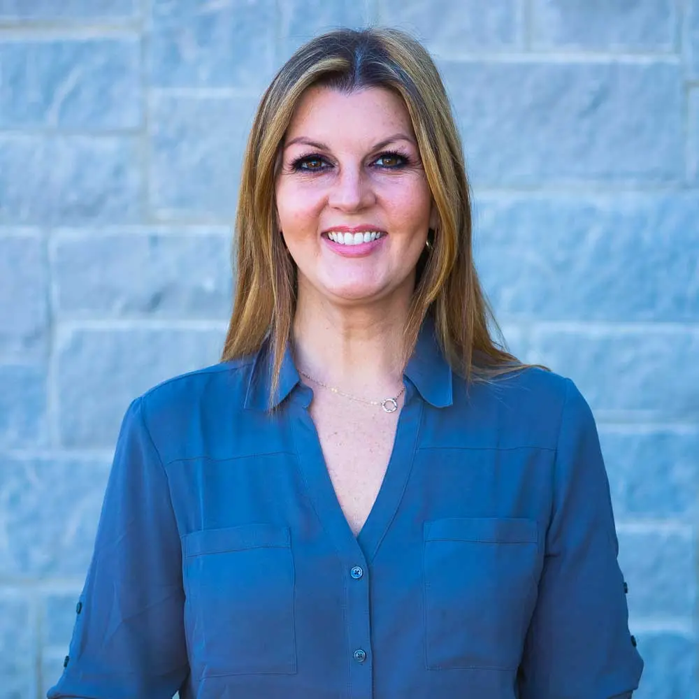 Woman with reddish-brown hair smiling at camera wearing dark blue button-up shirt against blue brick wall background