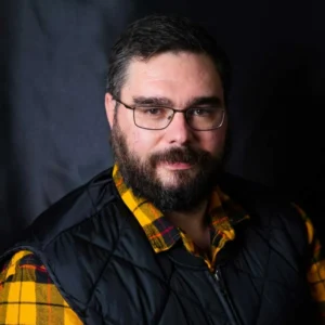 Man with dark hair, beard and glasses wearing a black and yellow plaid shirt, looking at camera against dark background