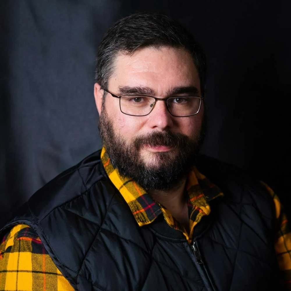 Man with dark hair, beard and glasses wearing a black and yellow plaid shirt, looking at camera against dark background