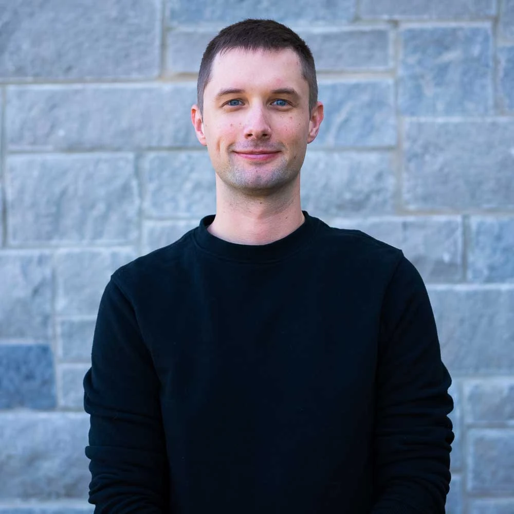 A young man with short dark hair wearing a black long-sleeved shirt against a stone brick wall background