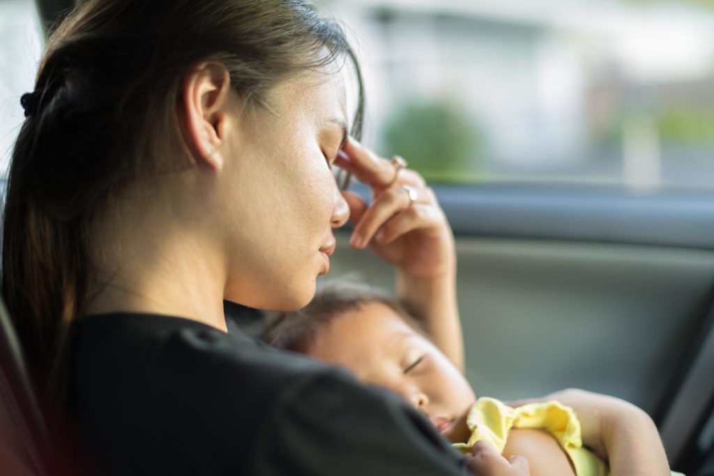 woman holding her infant while rubbing her forehead wondering what are the signs of postpartum depression in new mothers