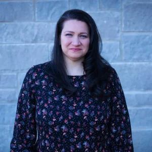 Woman with long dark hair wearing a dark blue floral patterned top smiling against a brick wall background