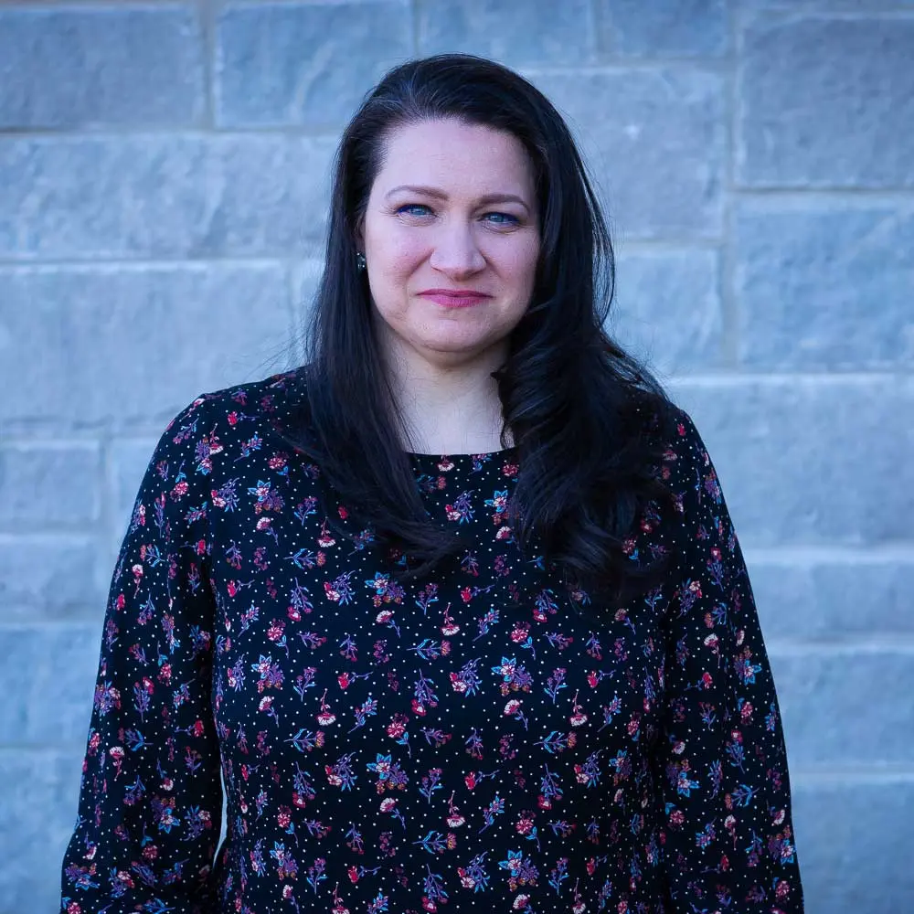 Woman with long dark hair wearing a dark blue floral patterned top smiling against a brick wall background