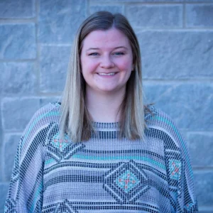 Woman with long blonde and gray hair wearing a patterned blue and white sweater, smiling at the camera against a brick wall