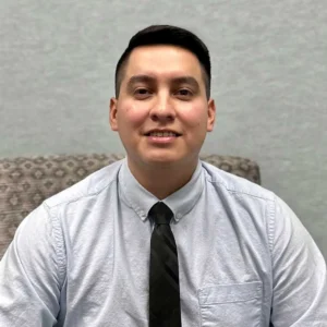 Man with short dark hair wearing a light gray button-up shirt and dark tie smiling at the camera indoors
