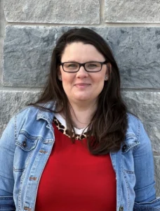 Woman with long dark hair and glasses wearing a denim jacket over a red top, smiling in front of a stone wall.