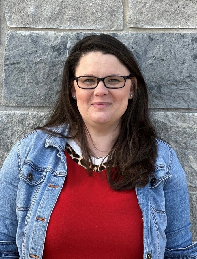 Woman with long dark hair and glasses wearing a denim jacket over a red top, smiling in front of a stone wall.