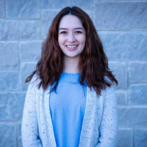 Smiling woman with long dark reddish-brown hair wearing blue dotted top and cream cardigan against brick wall