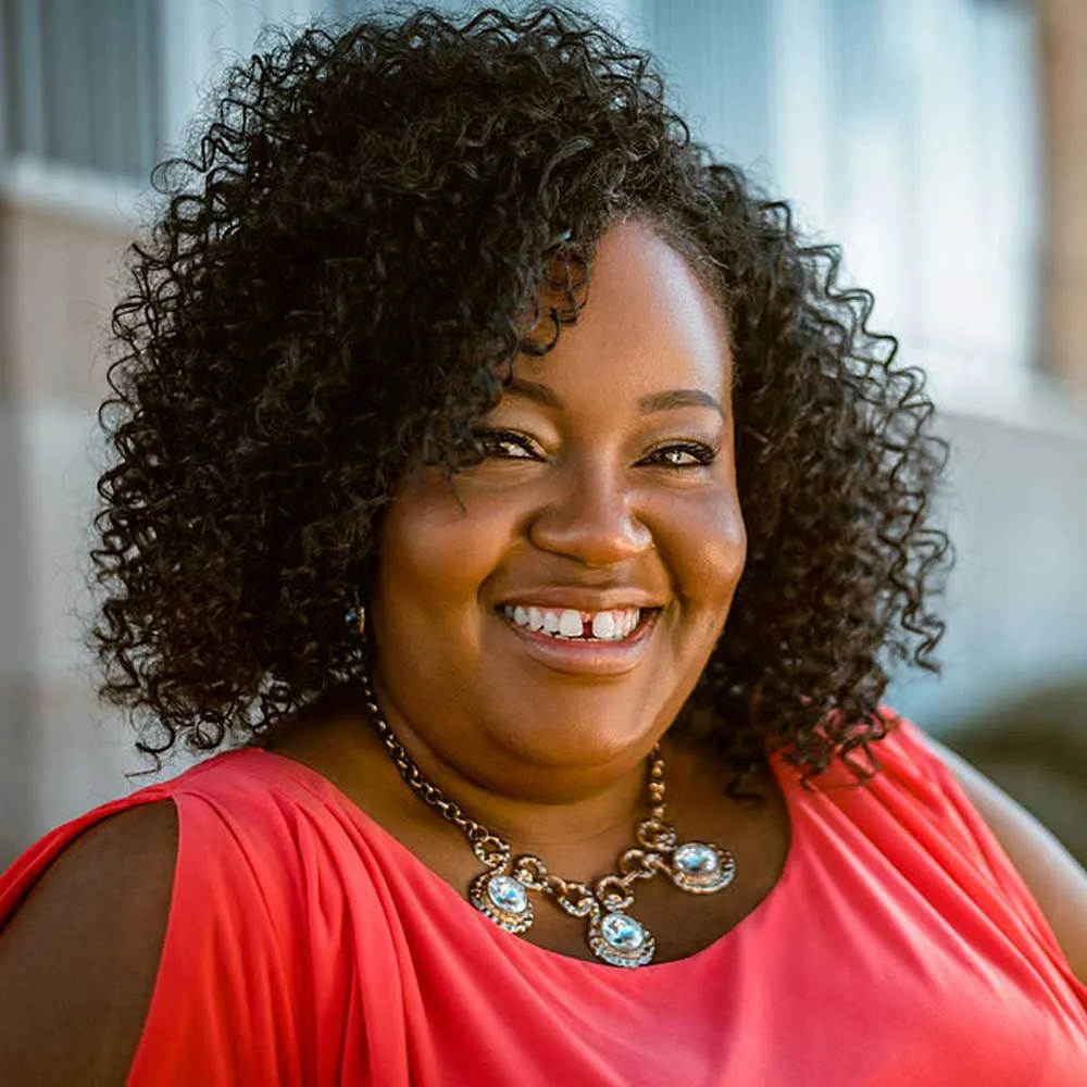 A woman with curly dark hair smiling warmly at the camera, wearing a bright red sleeveless top and decorative beaded necklace with blue stones