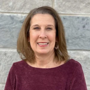 Woman with shoulder-length brown hair wearing maroon sweater, smiling at camera against brick wall