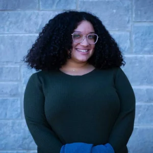 Woman with curly dark hair wearing glasses and a dark green ribbed sweater with blue fabric visible, smiling at camera.