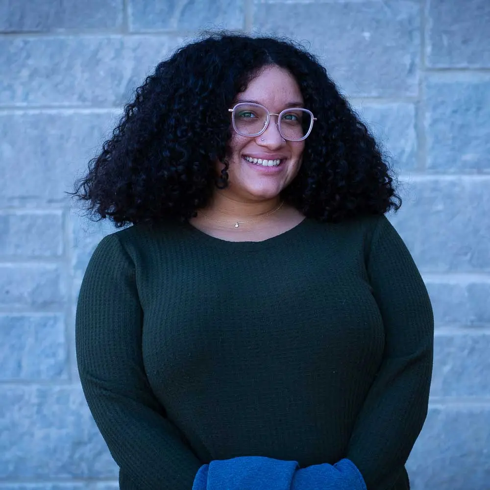 Woman with curly dark hair wearing glasses and a dark green ribbed sweater with blue fabric visible, smiling at camera.