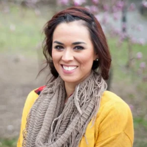 Woman with reddish-brown wavy hair wearing a bright yellow cardigan and tan scarf, smiling outdoors with purple flowers blurred in background