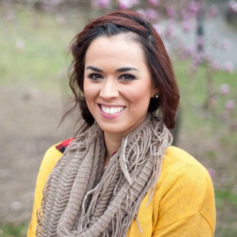 Woman with reddish-brown wavy hair wearing a bright yellow cardigan and tan scarf, smiling outdoors with purple flowers blurred in background