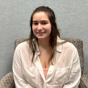 A young woman with long dark hair smiling at the camera, wearing a light pink button-up shirt over a coral top, seated against a gray background