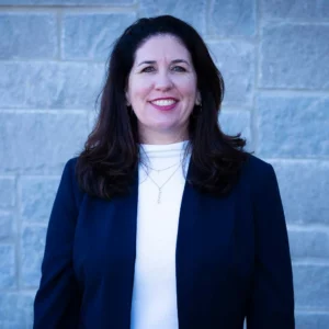Woman with long dark hair wearing dark blue blazer over white top, smiling at camera against brick wall