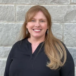 A woman with long reddish-blonde hair smiling at camera wearing a black zip-up top against a stone wall