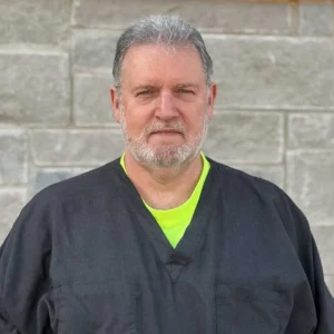 A man with gray hair and beard wearing a black shirt with a neon green undershirt against a stone wall