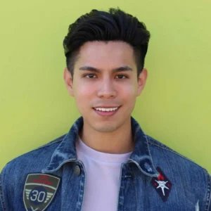 Man with dark styled hair wearing a denim jacket with patches over a light pink shirt against yellow background