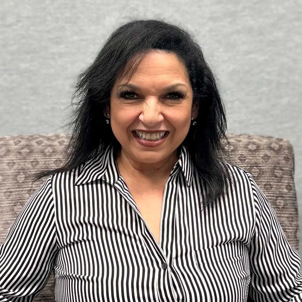 Woman with long black hair wearing a black and white striped shirt, smiling broadly at the camera, seated against a gray background.