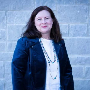 Woman with long dark hair wearing a dark blue denim jacket over a white top with a beaded necklace, smiling at camera against a brick background.