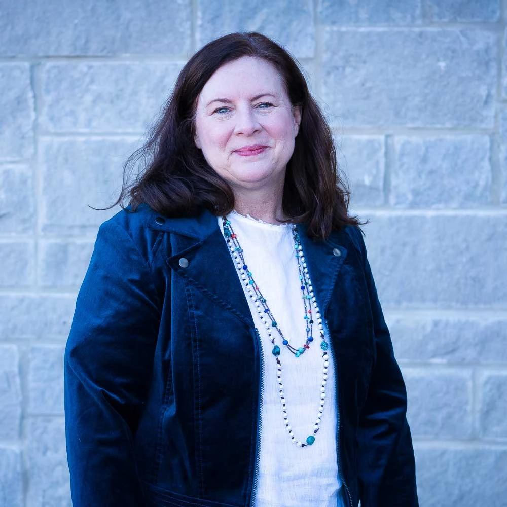 Woman with long dark hair wearing a dark blue denim jacket over a white top with a beaded necklace, smiling at camera against a brick background.