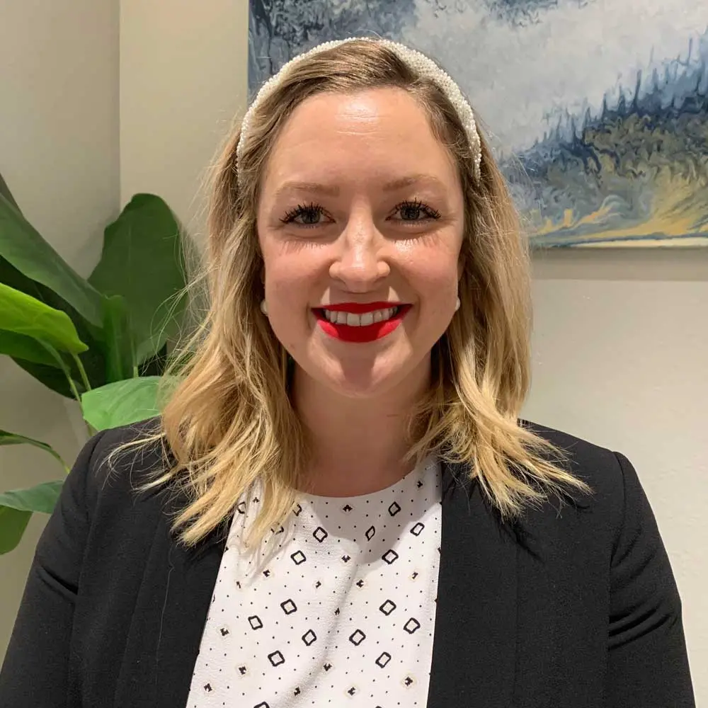Woman with blonde wavy hair wearing red lipstick, black blazer, and patterned white blouse, smiling with green plant visible