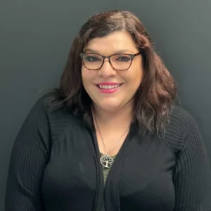 Woman with dark wavy hair and glasses wearing dark professional clothing with a cross pendant necklace, smiling at the camera