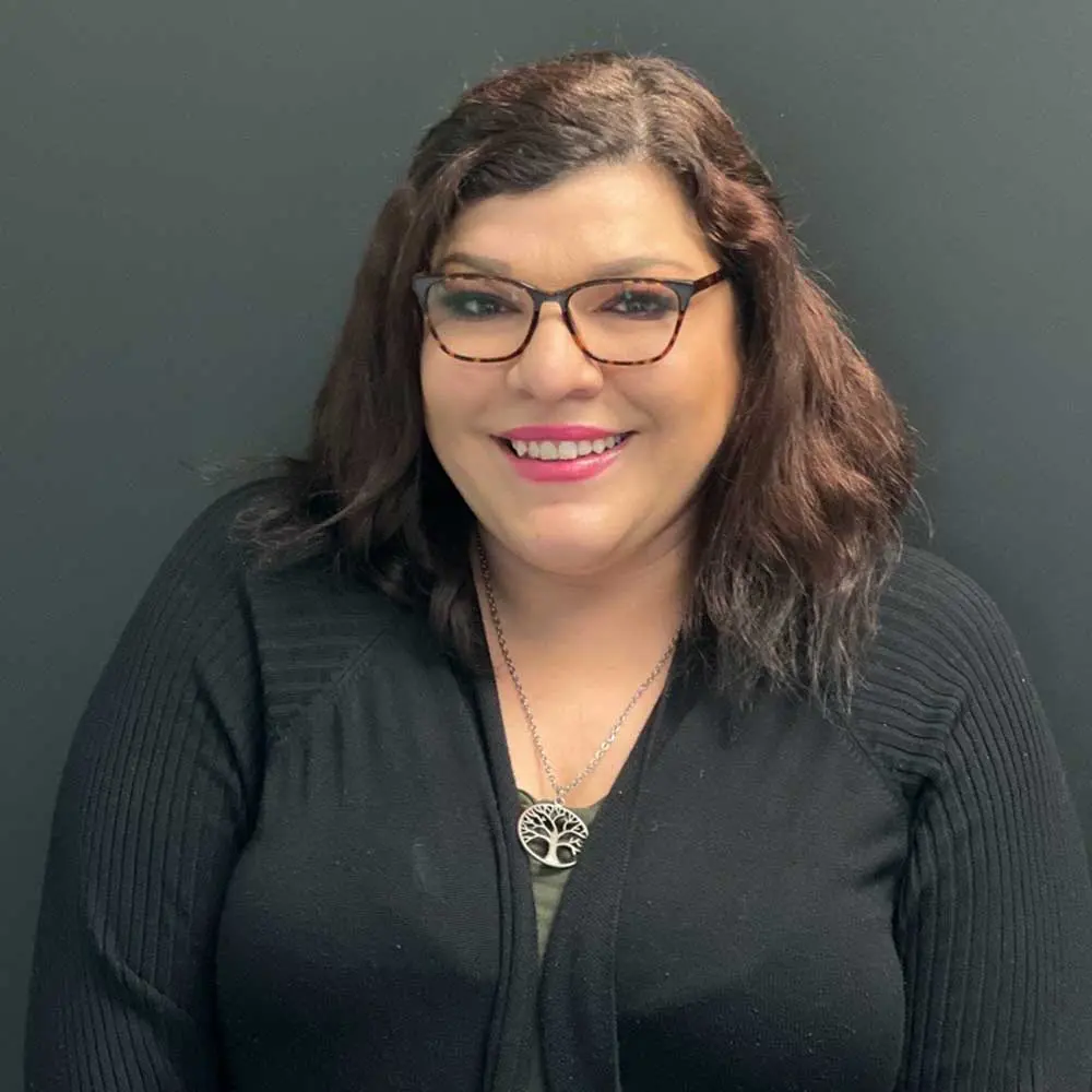 Woman with dark wavy hair and glasses wearing dark professional clothing with a cross pendant necklace, smiling at the camera