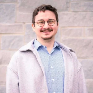 Man with dark hair and glasses wearing light pink cardigan over blue checkered shirt, smiling at camera against brick wall