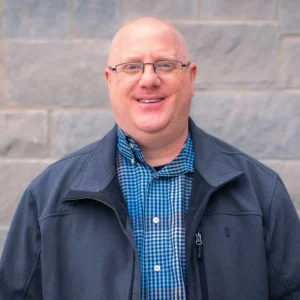 Man with glasses wearing blue checkered shirt and dark jacket, smiling at camera against brick background