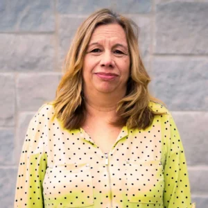Woman with shoulder-length wavy blonde hair wearing a chartreuse yellow shirt with small black dots, smiling at camera against a brick wall.