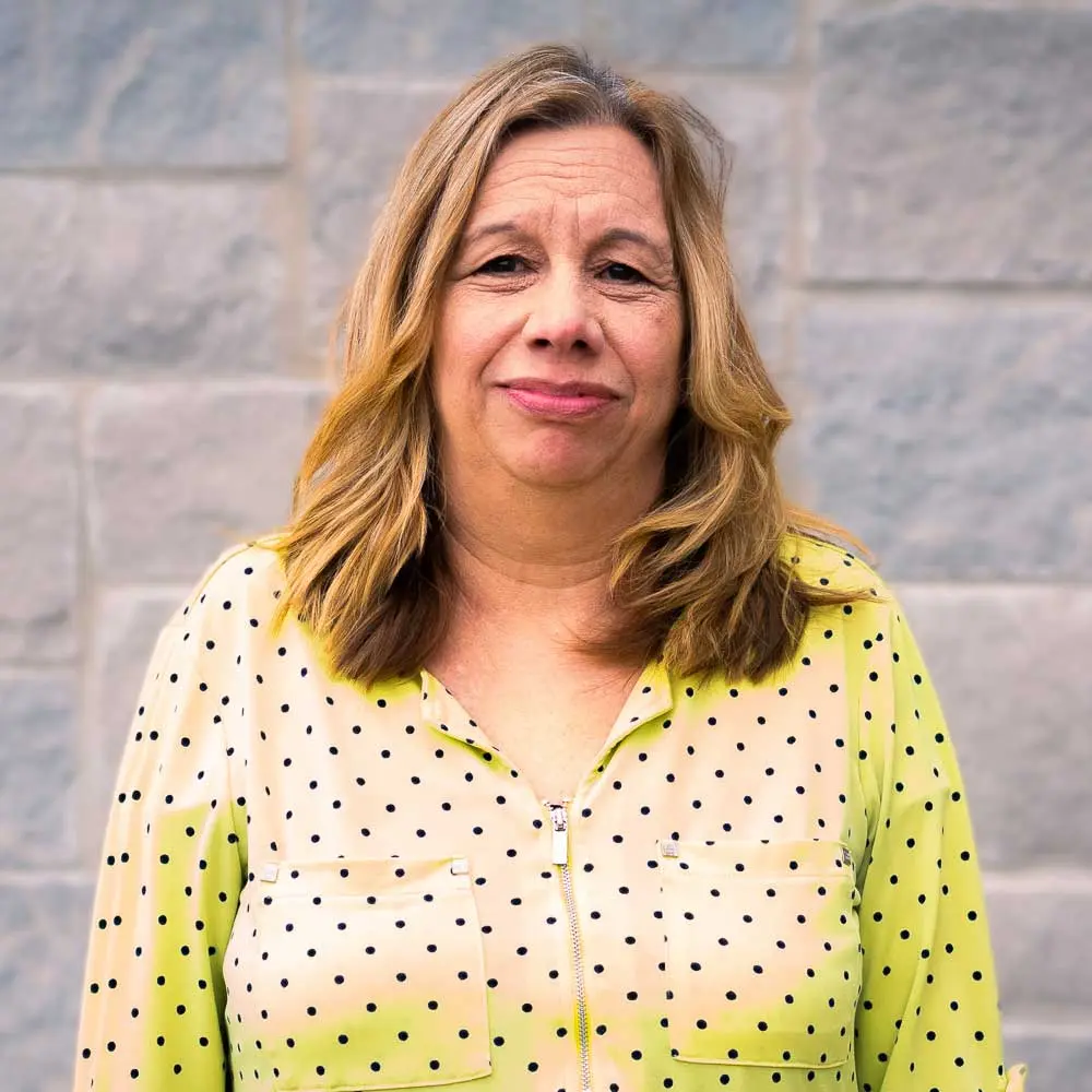 Woman with shoulder-length wavy blonde hair wearing a chartreuse yellow shirt with small black dots, smiling at camera against a brick wall.