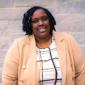 Professional headshot of a woman smiling at camera wearing a tan blazer over a white and blue plaid shirt against a brick wall background.