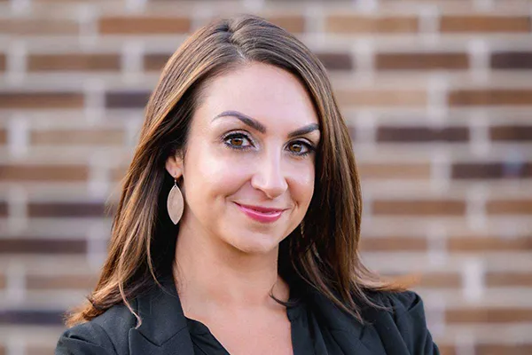 Headshot of woman with long brown hair and gold hoop earrings, wearing dark blazer against brick wall backdrop