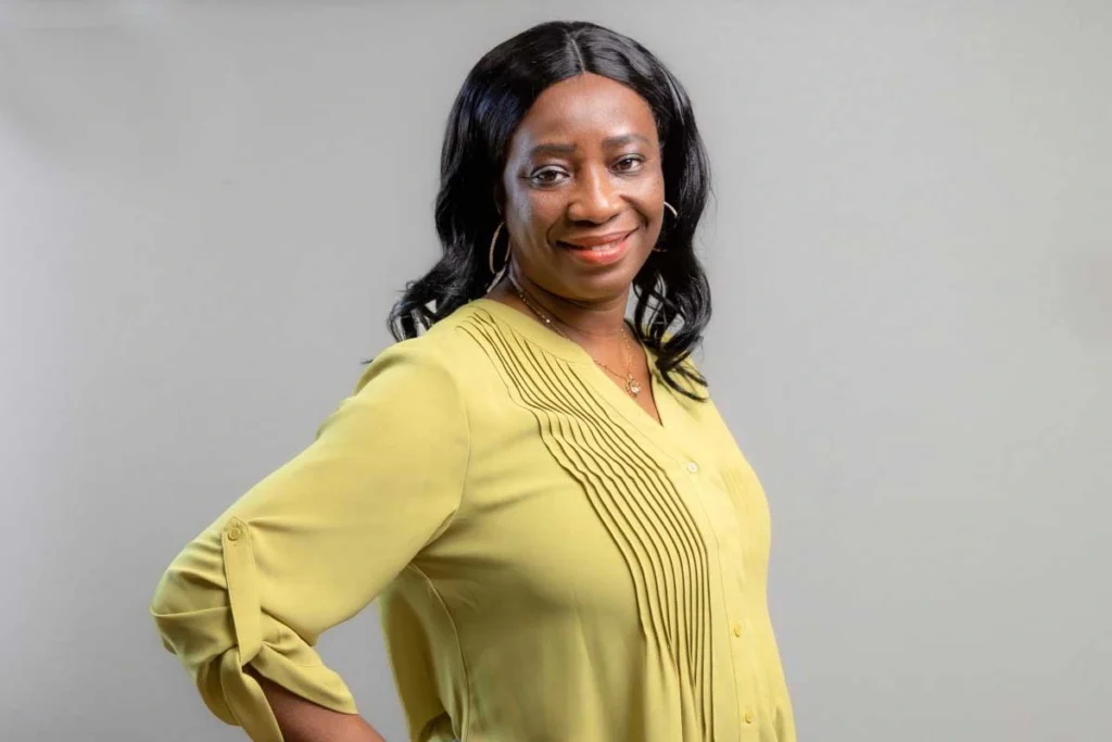 Woman smiling at camera wearing yellow pleated blouse with gold hoop earrings against gray background