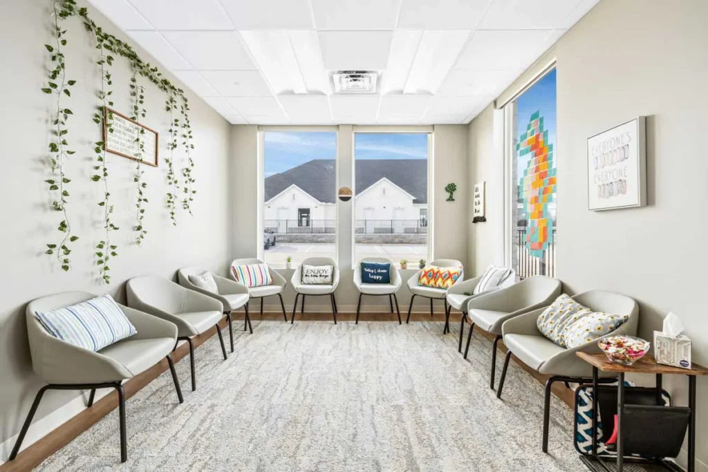 Bright modern therapy room with comfortable beige chairs arranged in circle, hanging green plants, large windows showing mountain views, and colorf...