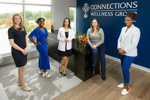 Five women standing together indoors in front of blue Connections Wellness Group sign with large window and flowers visible behind them