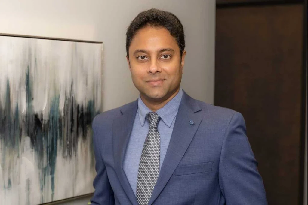 Man in blue dress shirt and patterned tie wearing business suit jacket, professional headshot against neutral background with abstract art