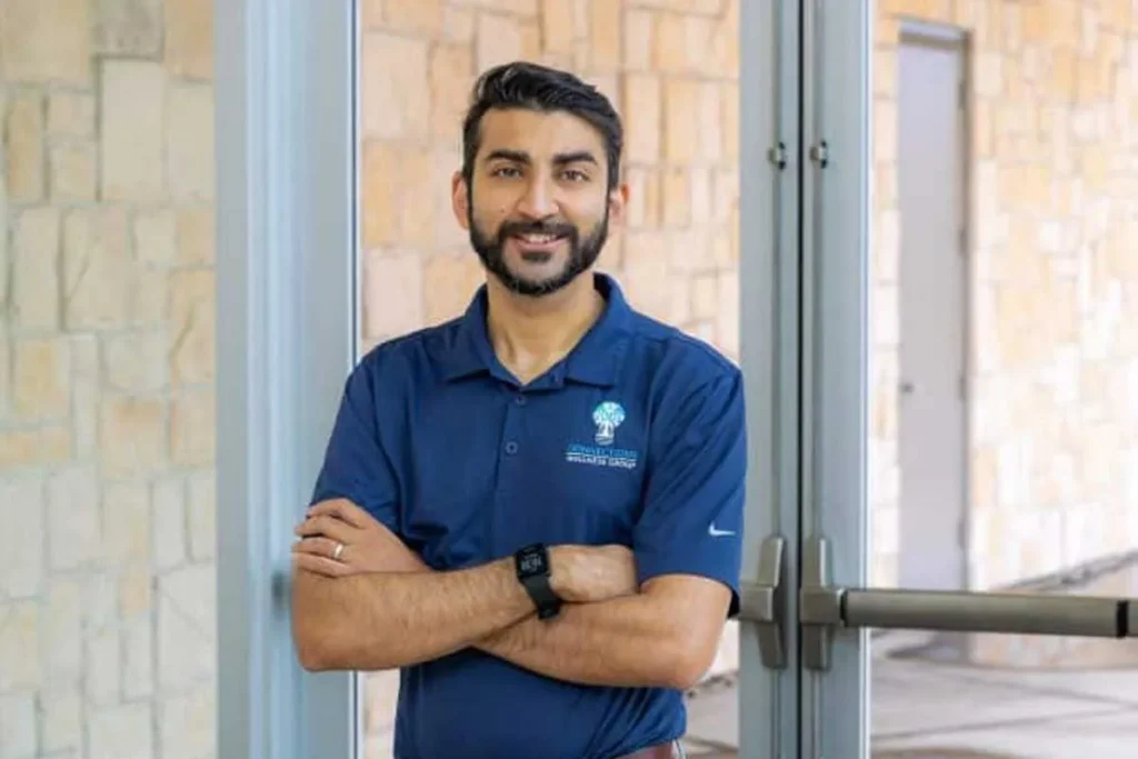 Man with dark hair and beard wearing blue polo shirt with logo, smiling at camera, arms crossed, brick wall background